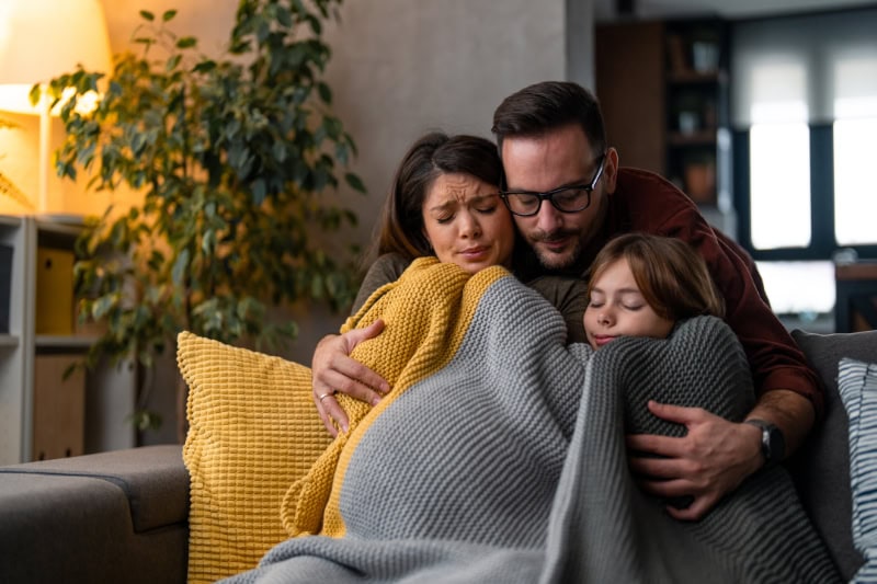 Why Isn’t My Furnace Heating My Entire Home? Photo of a family of three freezing in their home, huddled under a blanket to keep warm.