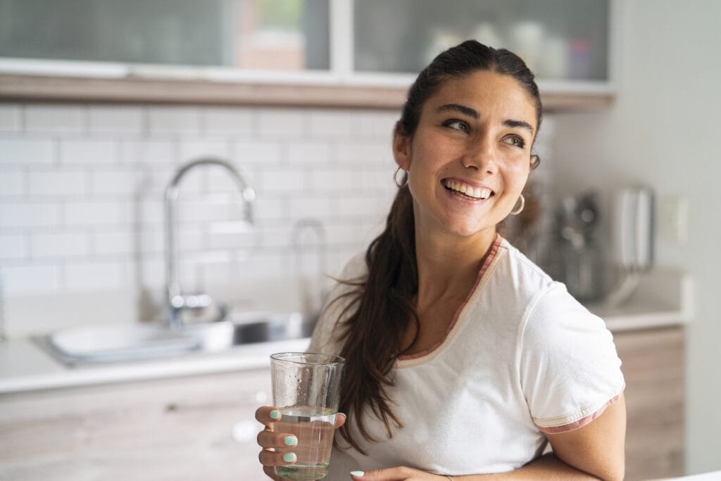 Top 10 Things Plumbers Wish You Were Privy To. Woman smiling holding a glass of water in her kitchen.